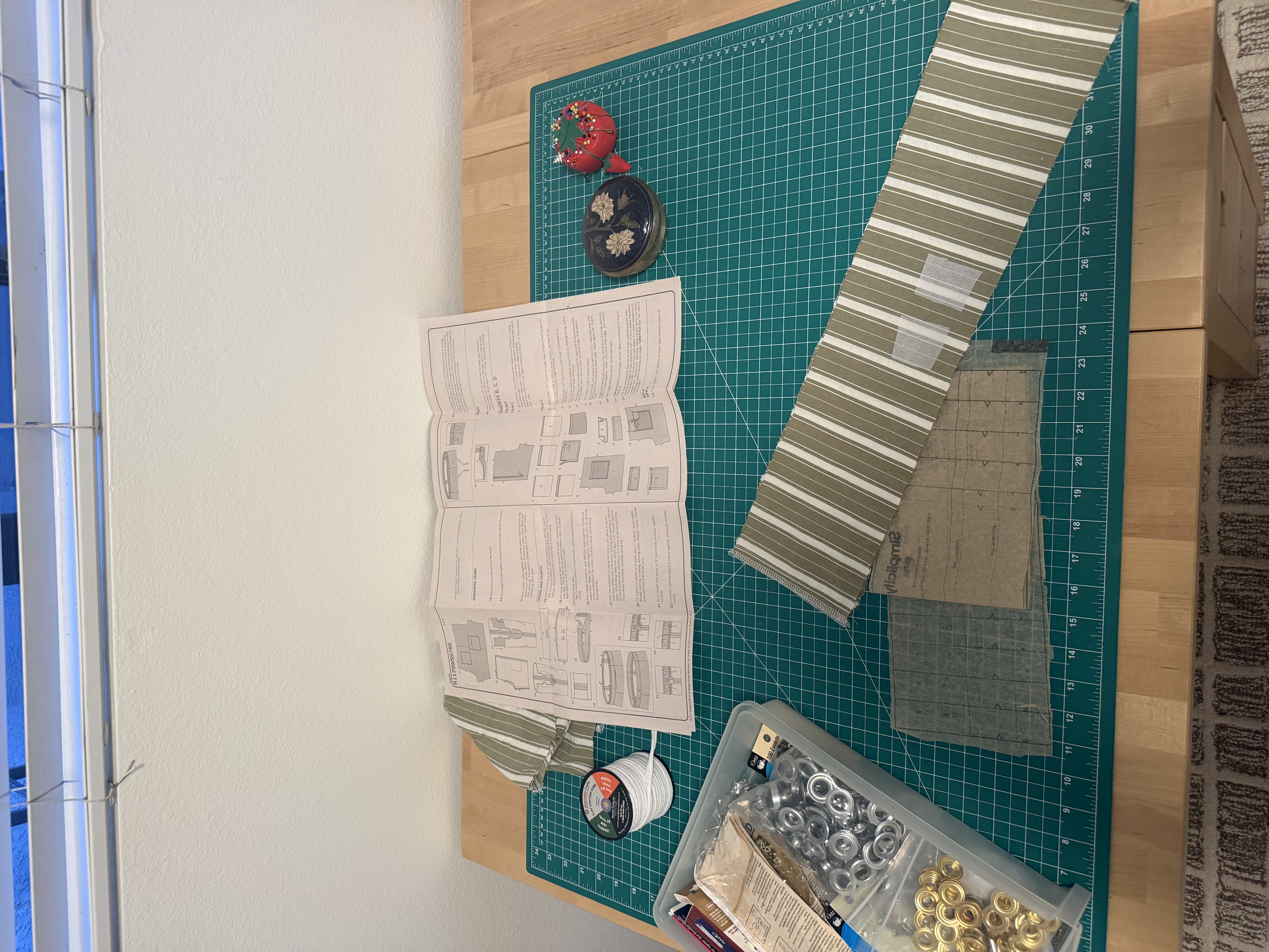 Grommet hardware, drawstring cord, and the Simplicity pattern instructions laid out on the cutting mat